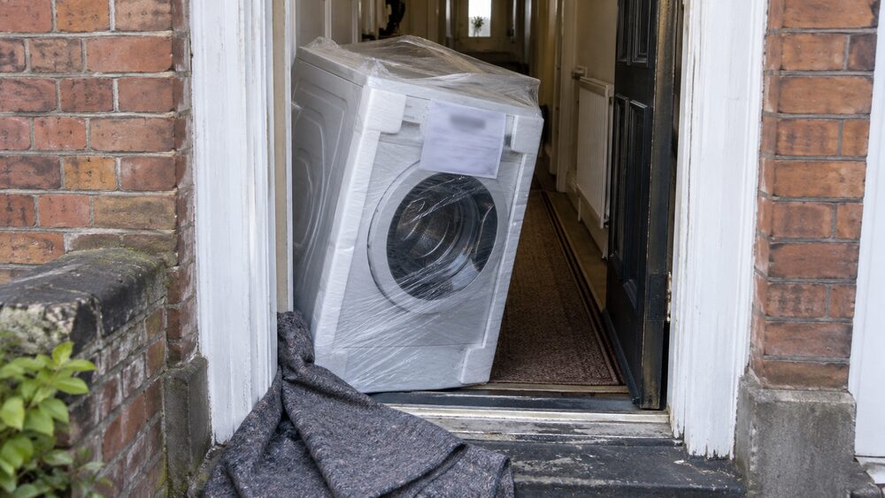 A washing machine wrapped in film is tilted through the front door of a terraced house.