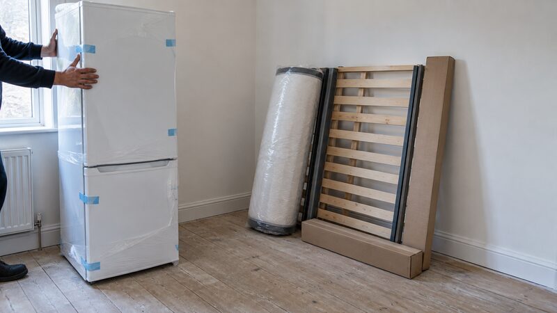 A new fridge and a flat-packed bed in an empty room with bare floorboards.