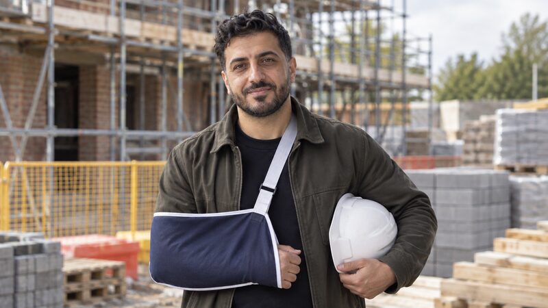 A man stands on a building site with his arm in a sling and a hard hat under his other arm.