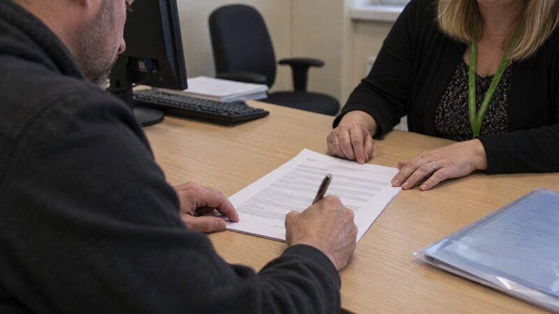 A hand signs a printed form on a desk while another hand rests near the page.