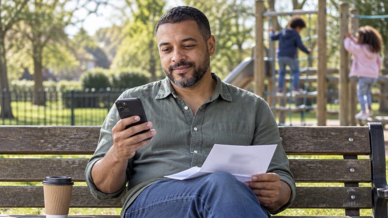 A man sits on a park bench reading a letter while children play behind him.
