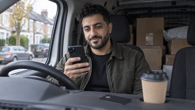 A man sits in his parked van checking his phone with a card on the dashboard.