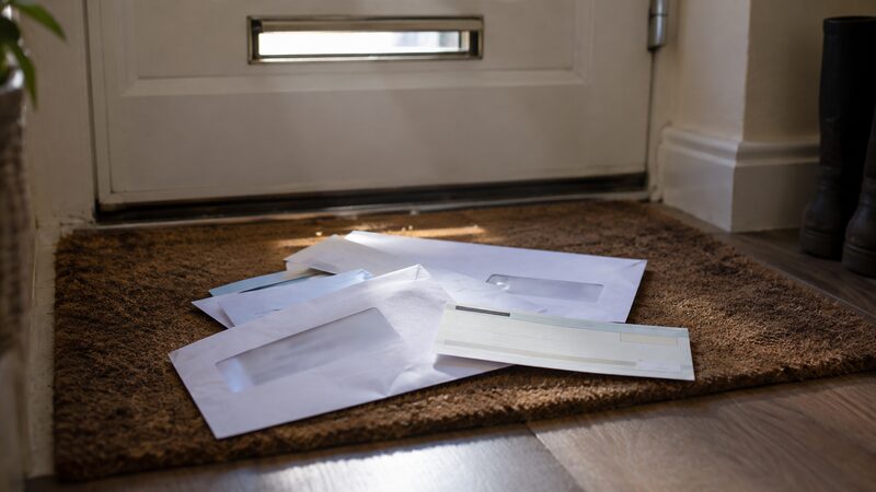 Pile of unopened envelopes and cheque-style slips lying on a doormat by a front door.