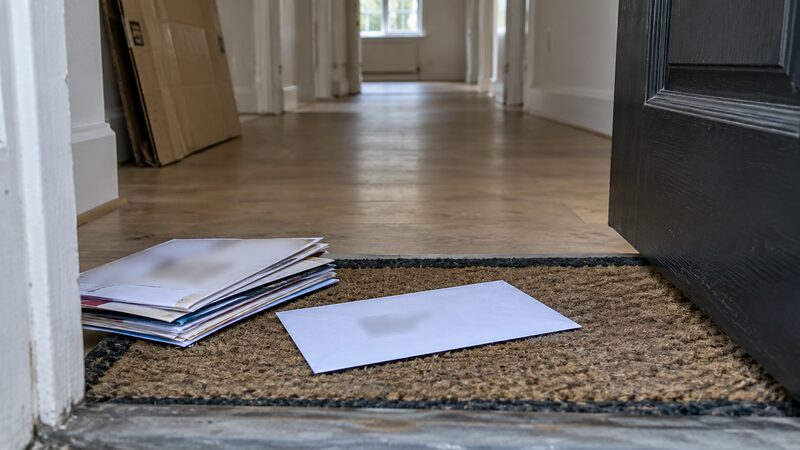 A plain envelope lies on a doormat inside a front door with uncollected post beside it.