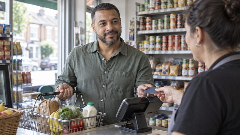 A man holds a shopping basket at a shop till after tapping his debit card.