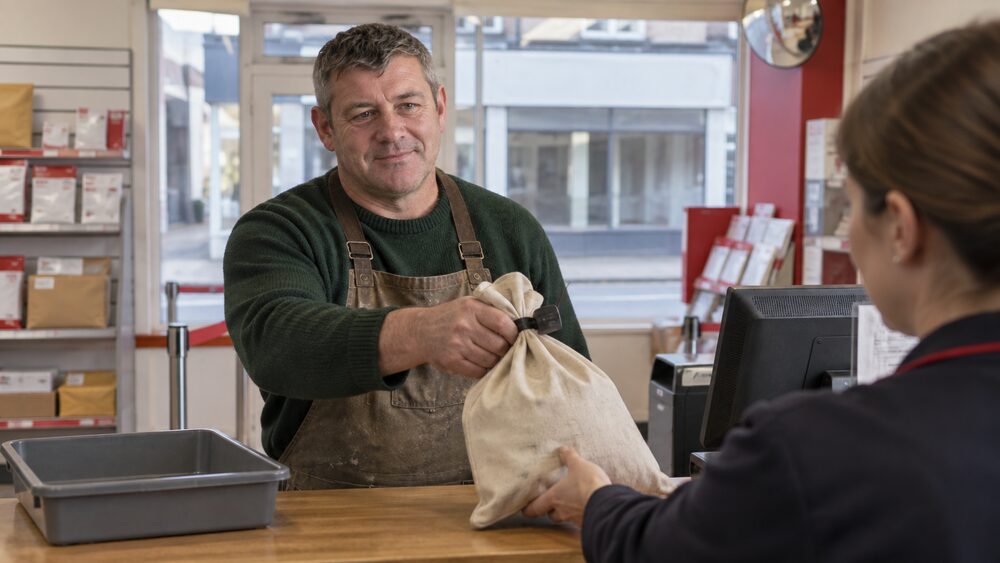 A tradesman in a work apron hands a sealed deposit bag to a clerk at a post office counter.