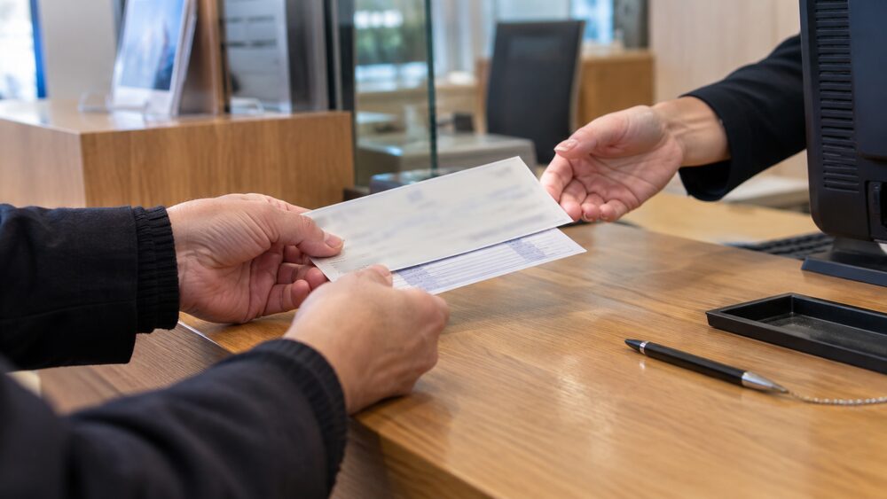 Hands pass a cheque and paying-in slip across a bank counter.