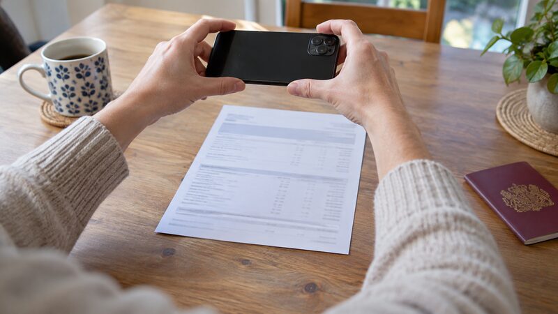 Hands hold a smartphone above a bank statement on a table, with a passport nearby.