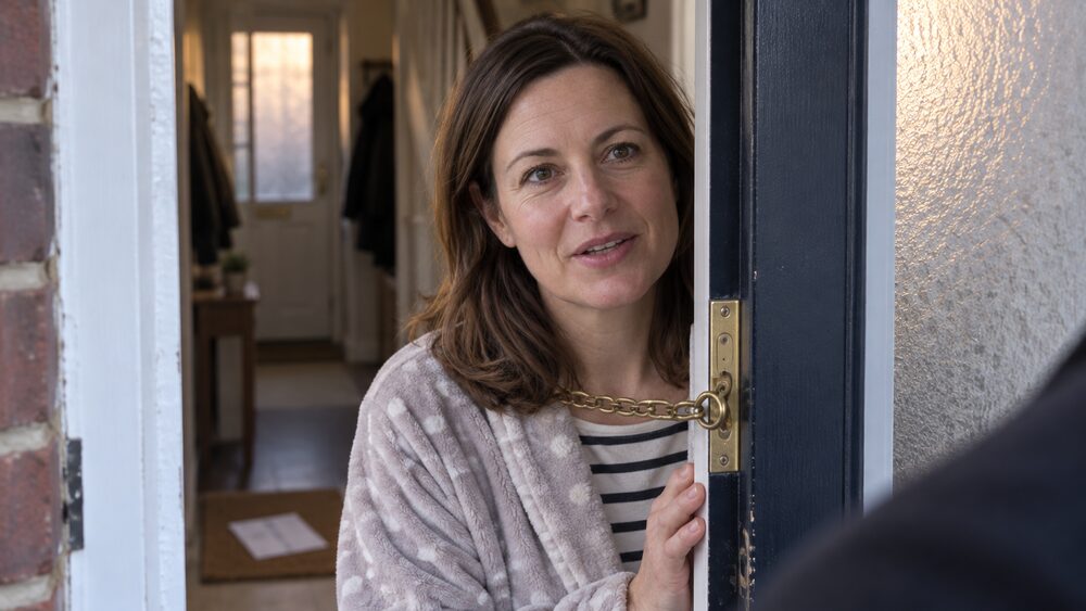 A woman in a dressing gown speaks through a chained front door with a letter on the mat.