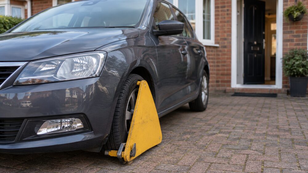 A car on a driveway has a wheel clamp fitted, with the house door slightly open behind.