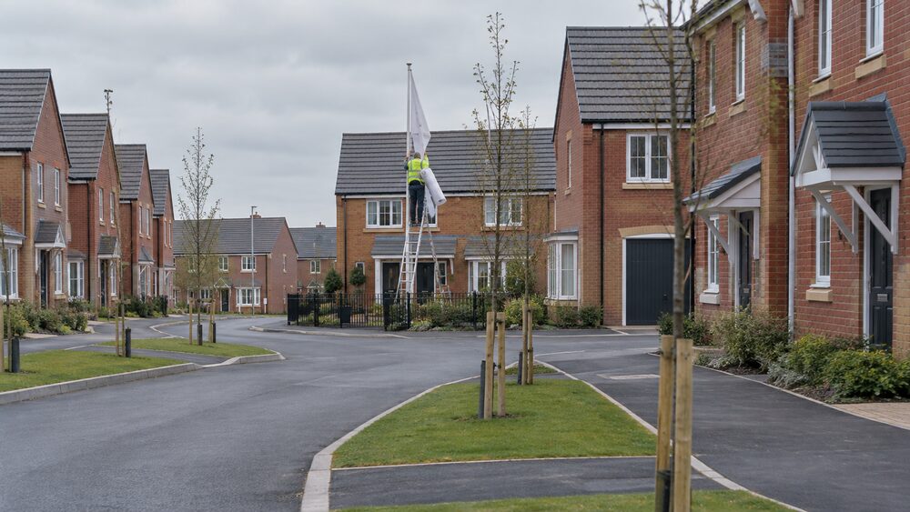 Rows of identical new brick houses on an overcast estate with saplings and a workman at the show home.