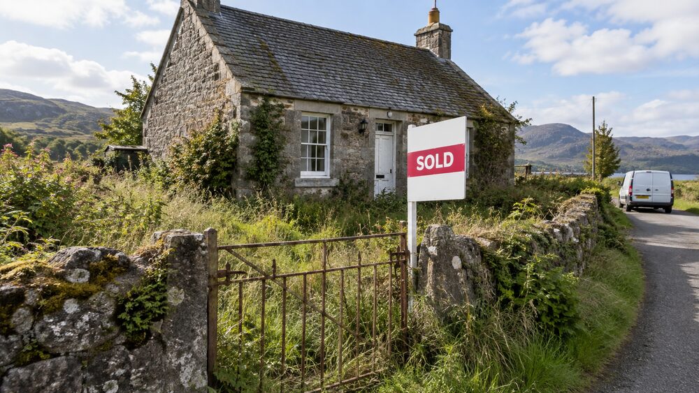 A small stone cottage with an overgrown garden and a sold board at the gate on a rural lane.