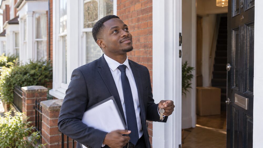 A young man holding keys and papers stands on the doorstep of a terraced house.