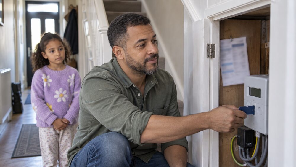 A man crouches at a prepayment meter with a top-up key while a child watches from the hallway.