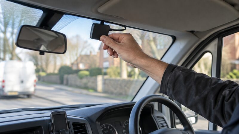 A hand fits a small telematics device to the inside of a van windscreen.