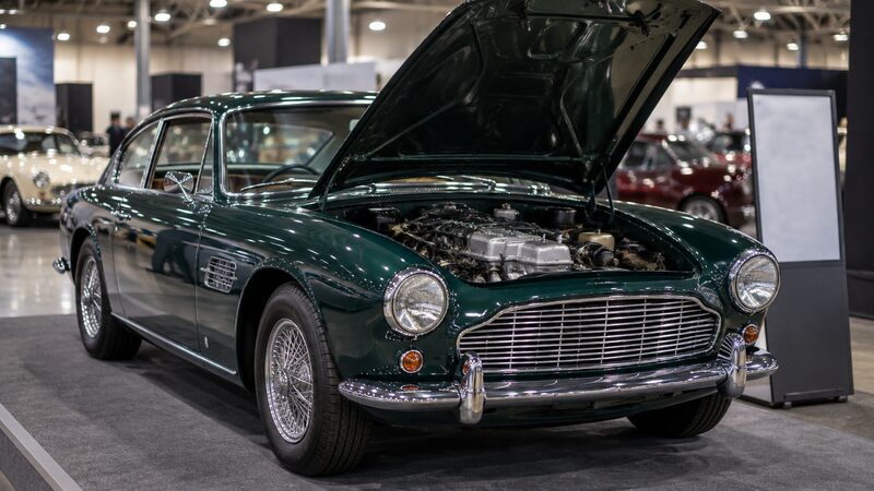A polished classic car with its bonnet open stands on a display stand at an indoor show.