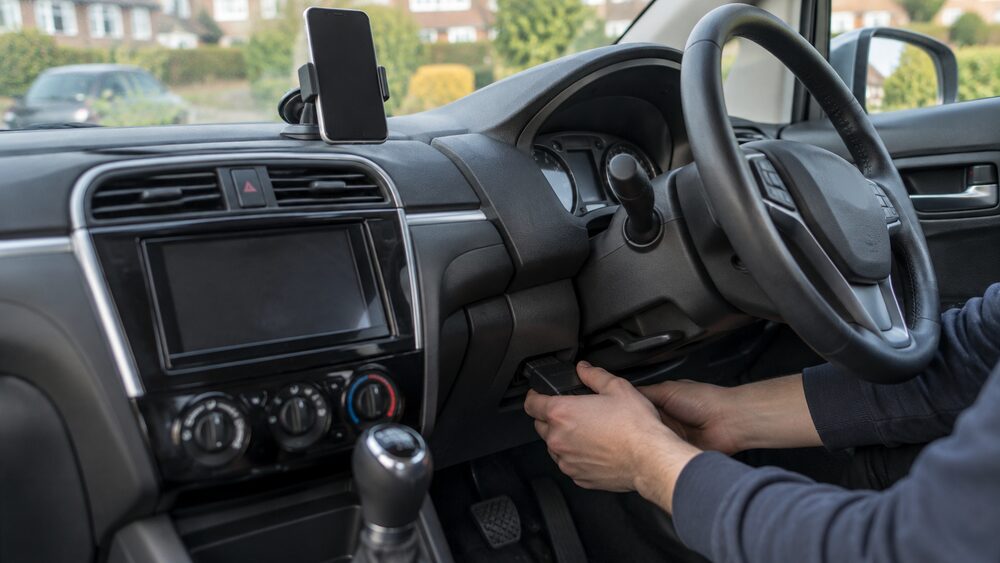 Hands plug a small device into a port under a car dashboard.