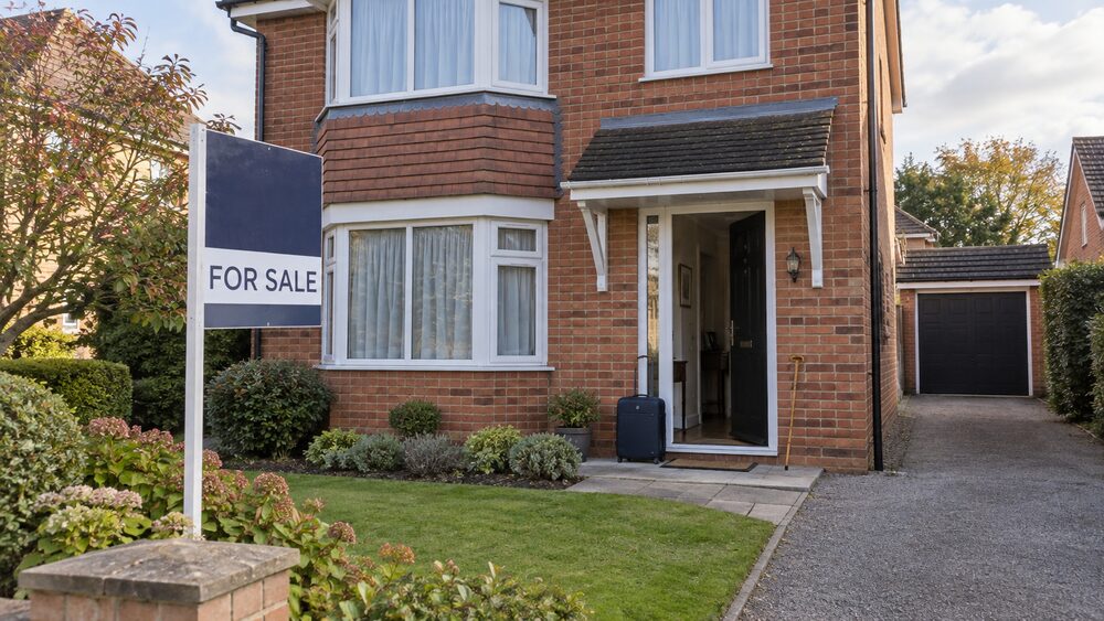 A detached brick house with a for sale board and a suitcase and walking stick by the open front door.