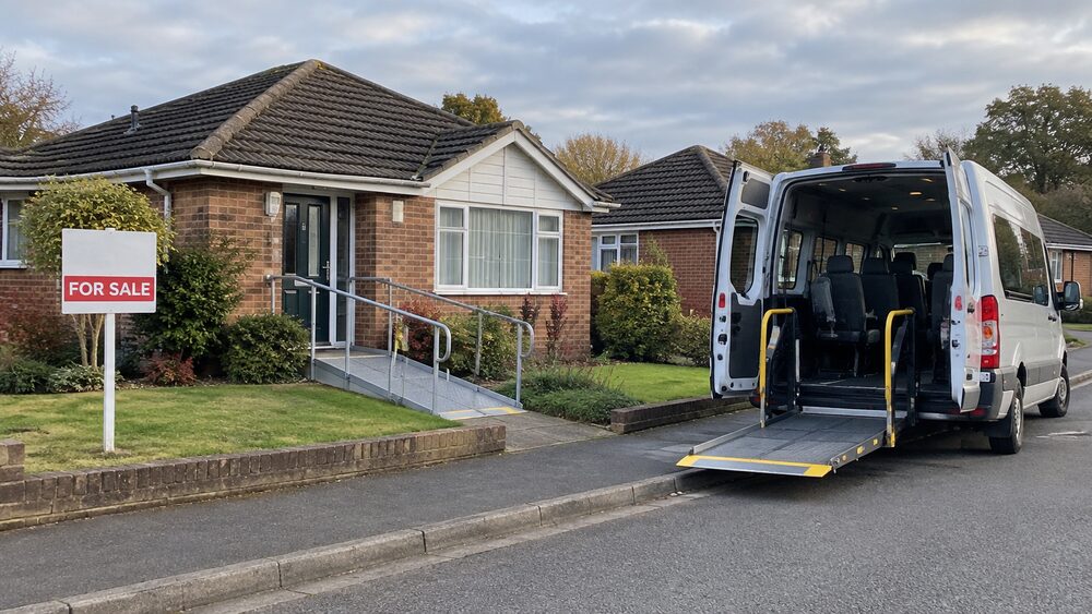 A bungalow with a wheelchair ramp and a for sale board, with a care home minibus waiting at the kerb.