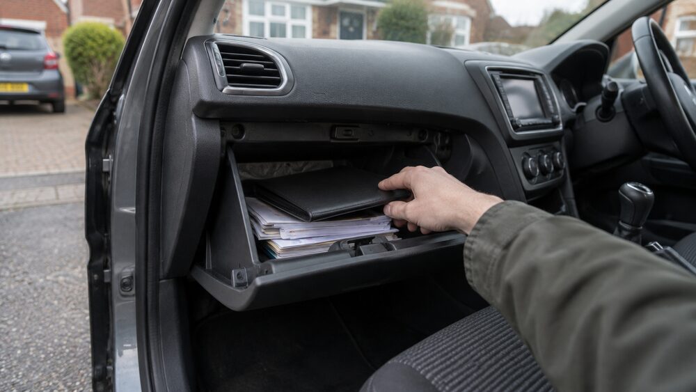 A hand reaches into an open glovebox toward a document wallet inside a car.
