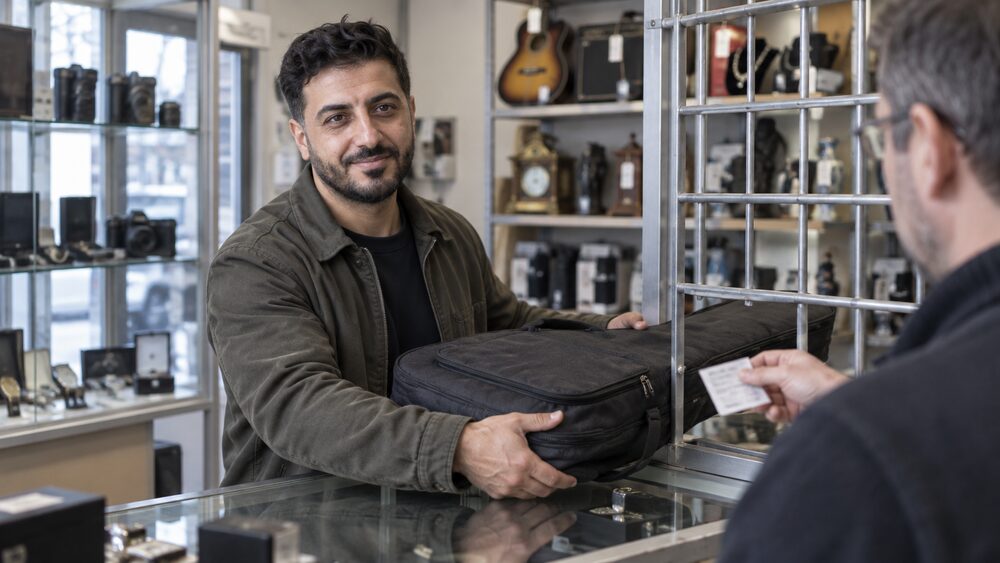 A man hands a guitar in a soft case to an assistant at a pawnbroker's counter.