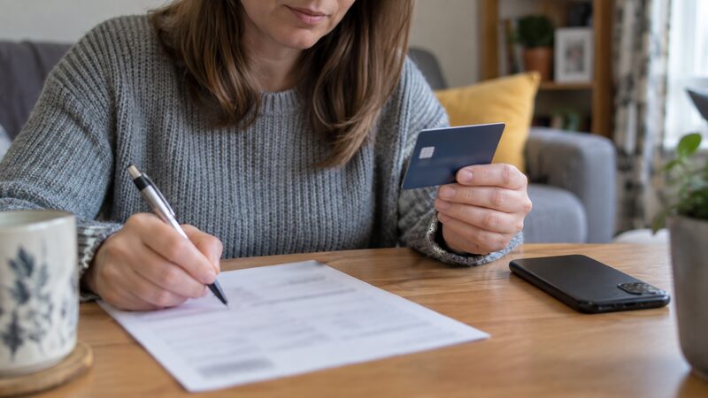A hand holds a debit card while another writes on a form at a desk.