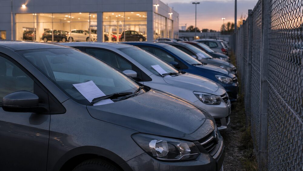 A row of handed-back hatchbacks parked by a fence behind a dealership at dusk.