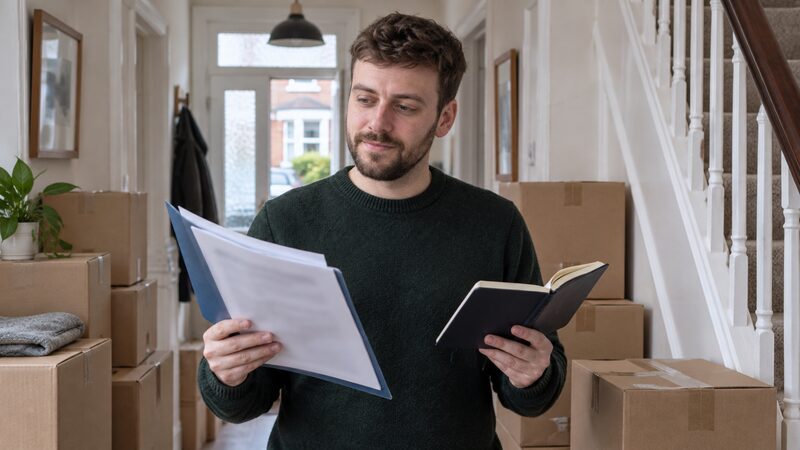 A man stands in a hallway with moving boxes, holding a folder and a diary.