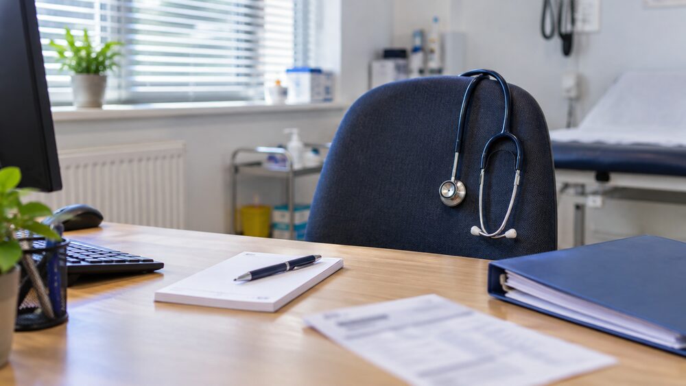 Stethoscope on chair and desk with prescription pad, payslip and policy folder in a consulting room.