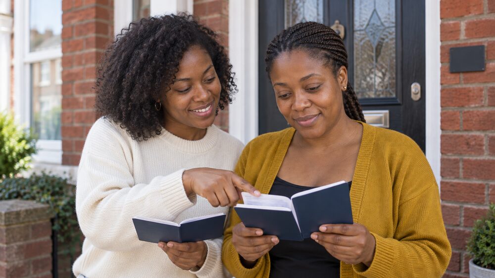 Two women compare open savings passbooks on the doorstep of a terraced house.