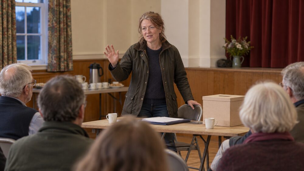 A woman stands to speak at a village hall meeting beside a ballot box and papers.