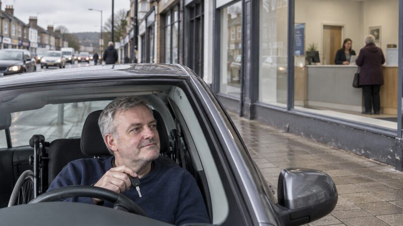 A wheelchair user sits in an adapted car at the kerb, looking towards a banking hub.