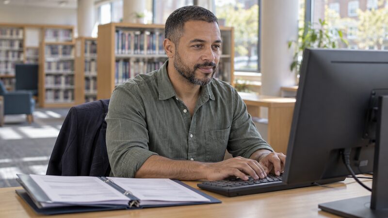 A man sits at a library computer desk filling in an online form with bank statements beside him.