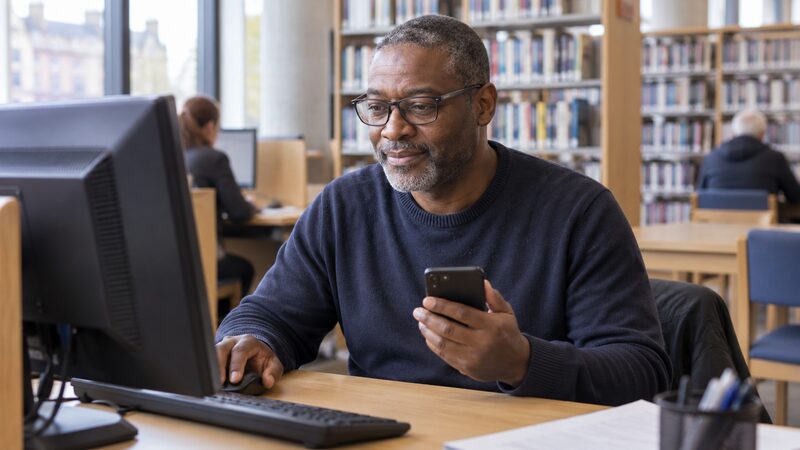 A man sits at a library computer, holding a phone and using the mouse.