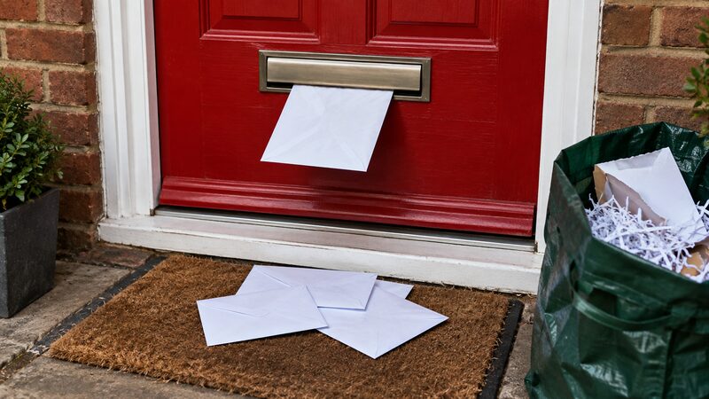 Envelopes spill from a red letterbox onto a doormat beside a recycling bag.