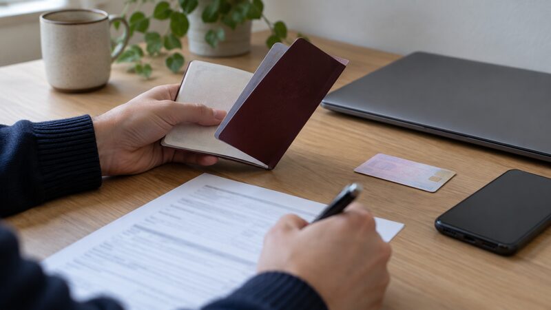 Hands at a desk hold a passport and write on a form beside a laptop.